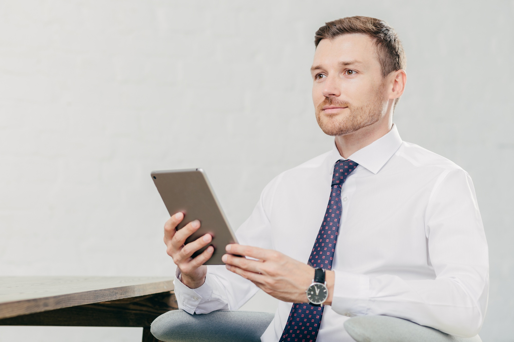 Thoughtful Caucasian male laywer thinks about something, holds modern tablet computer